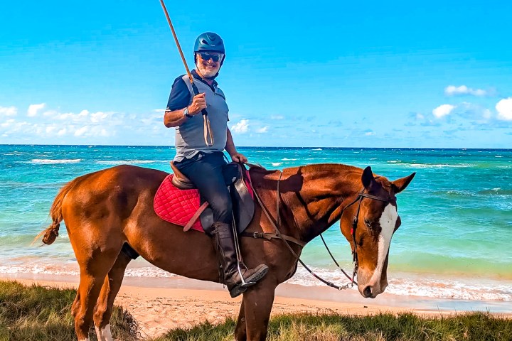 Person on horseback holding a pole on a beach, wearing a helmet under a clear blue sky.