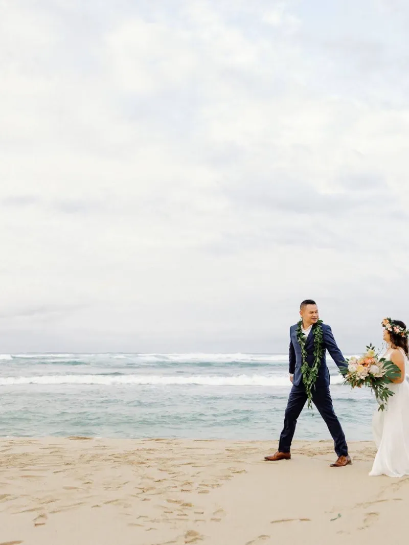 Bride and groom holding hands on a beach with ocean waves and cloudy sky.