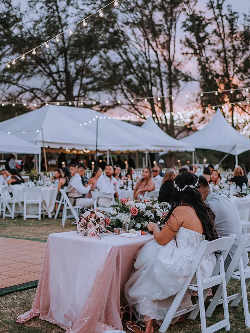 Outdoor wedding reception at sunset with guests seated under string lights.