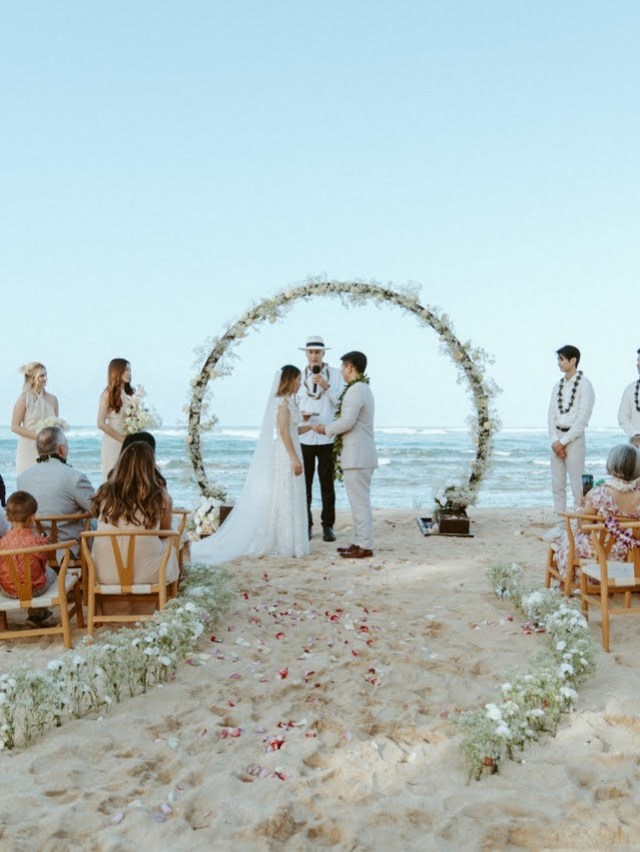 Beach wedding ceremony with arch, couple, guests, and sea in the background.