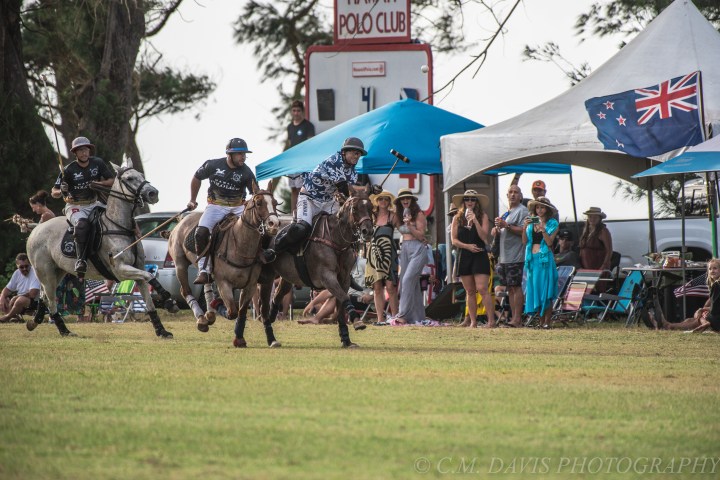 Polo match with players on horses, spectators, and a scoreboard in the background.