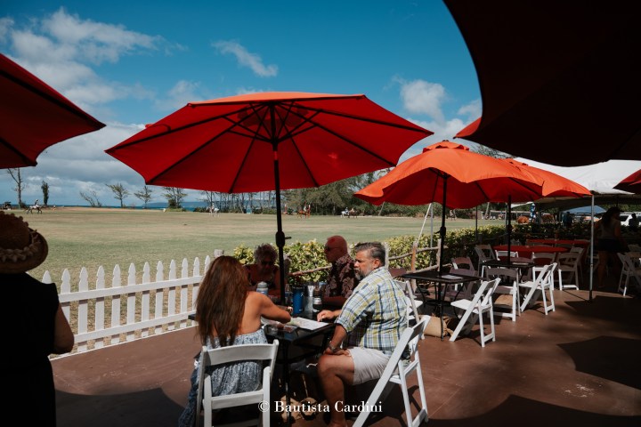 People seated under red umbrellas at an outdoor cafe with a grassy field view.