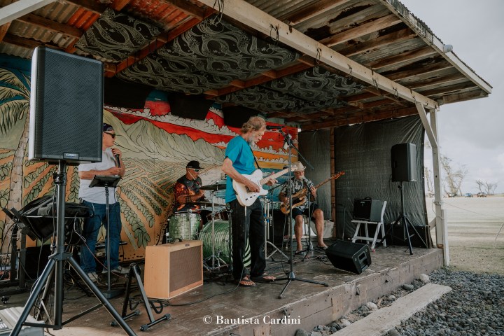 Five musicians performing on a wooden stage with a tropical mural backdrop.