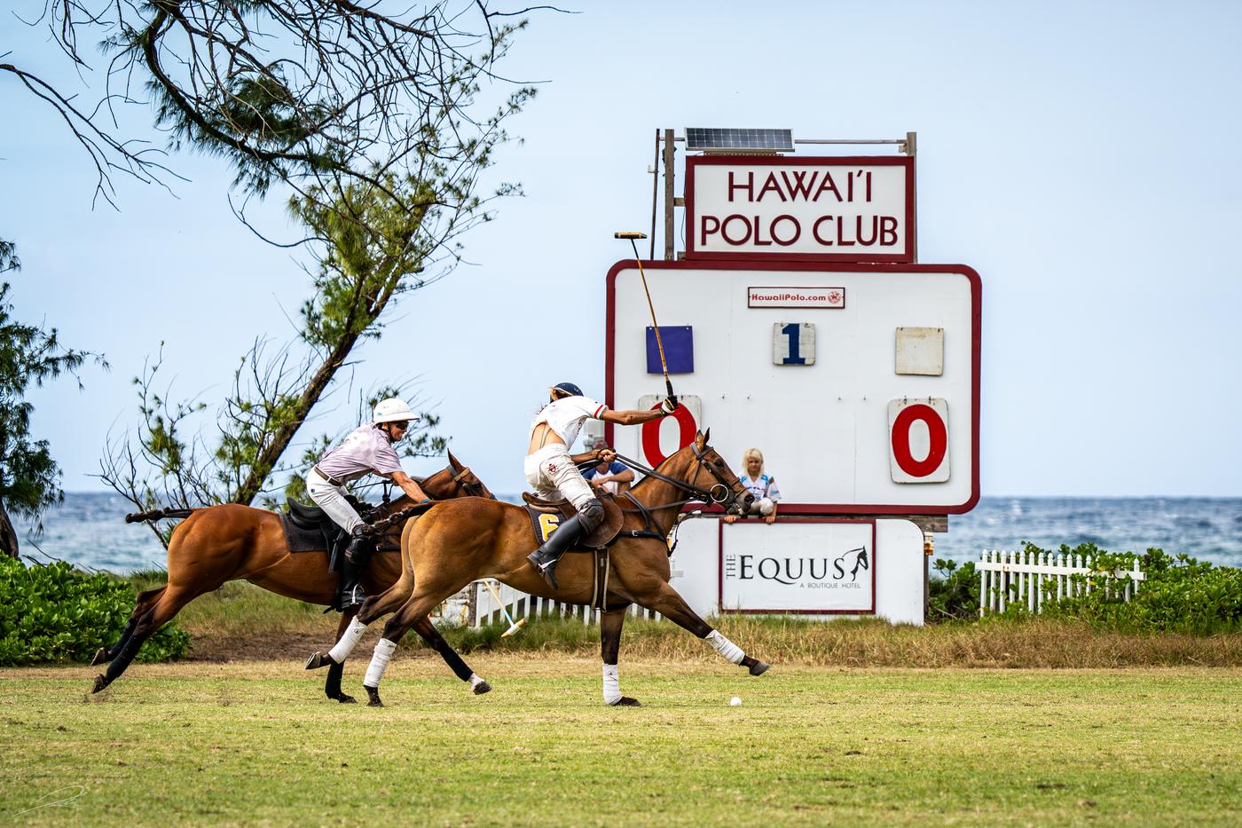 Two players ride off in front of the Hawaii Polo Club scoreboard with the Pacific behind